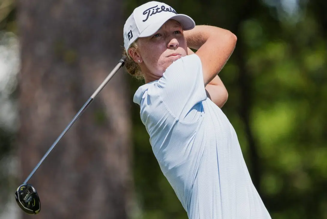 Emma McMyler wearing a light blue striped polo, khaki shorts, and a white Titleist hat, looking down the fairway while holding the follow-through of a golf swing with a driver against a background of blurred trees.