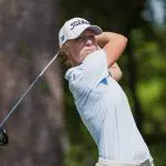 Emma McMyler wearing a light blue striped polo, khaki shorts, and a white Titleist hat, looking down the fairway while holding the follow-through of a golf swing with a driver against a background of blurred trees.