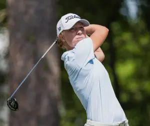 Emma McMyler wearing a light blue striped polo, khaki shorts, and a white Titleist hat, looking down the fairway while holding the follow-through of a golf swing with a driver against a background of blurred trees.