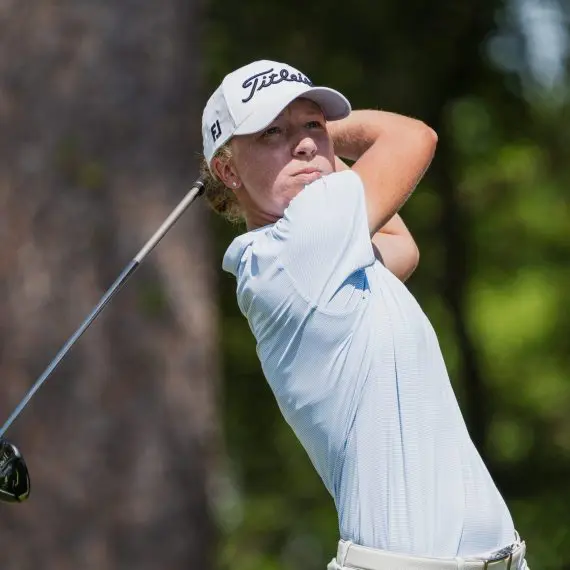 Emma McMyler wearing a light blue striped polo, khaki shorts, and a white Titleist hat, looking down the fairway while holding the follow-through of a golf swing with a driver against a background of blurred trees.